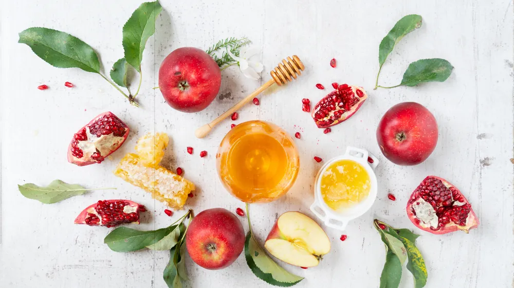 Apples, open pomegranates, a jar of honey and a honeycomb on a white table: the symbols of the Rosh Hashanah table