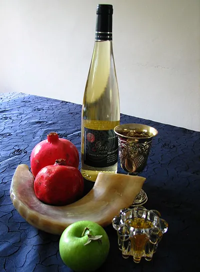 A ram's-horn shofar holding two pomegranates, with a green apple, a bottle of wine, a kiddush cup and a small dish of honey