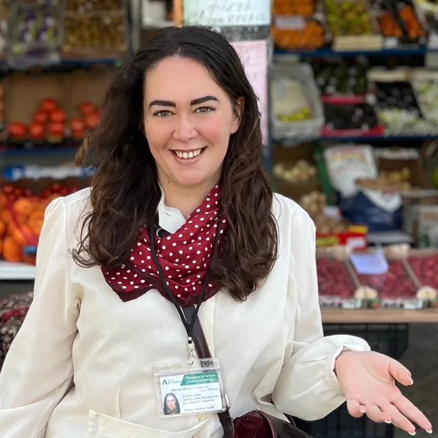 Clara, official tourism guide, in front of a fruit stall at a market, wearing her guide badge